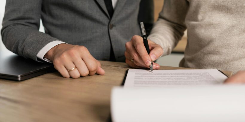 Close-up of businessmen signing documents at a wooden table in an office.