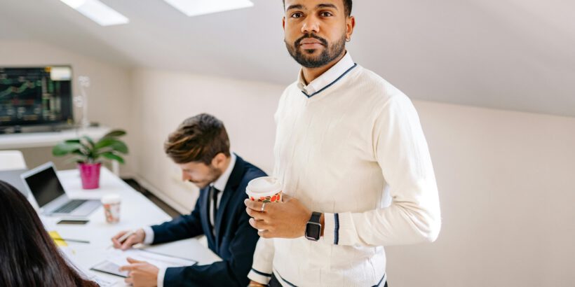 Confident businessman with coffee in a modern office setting with colleagues working nearby.